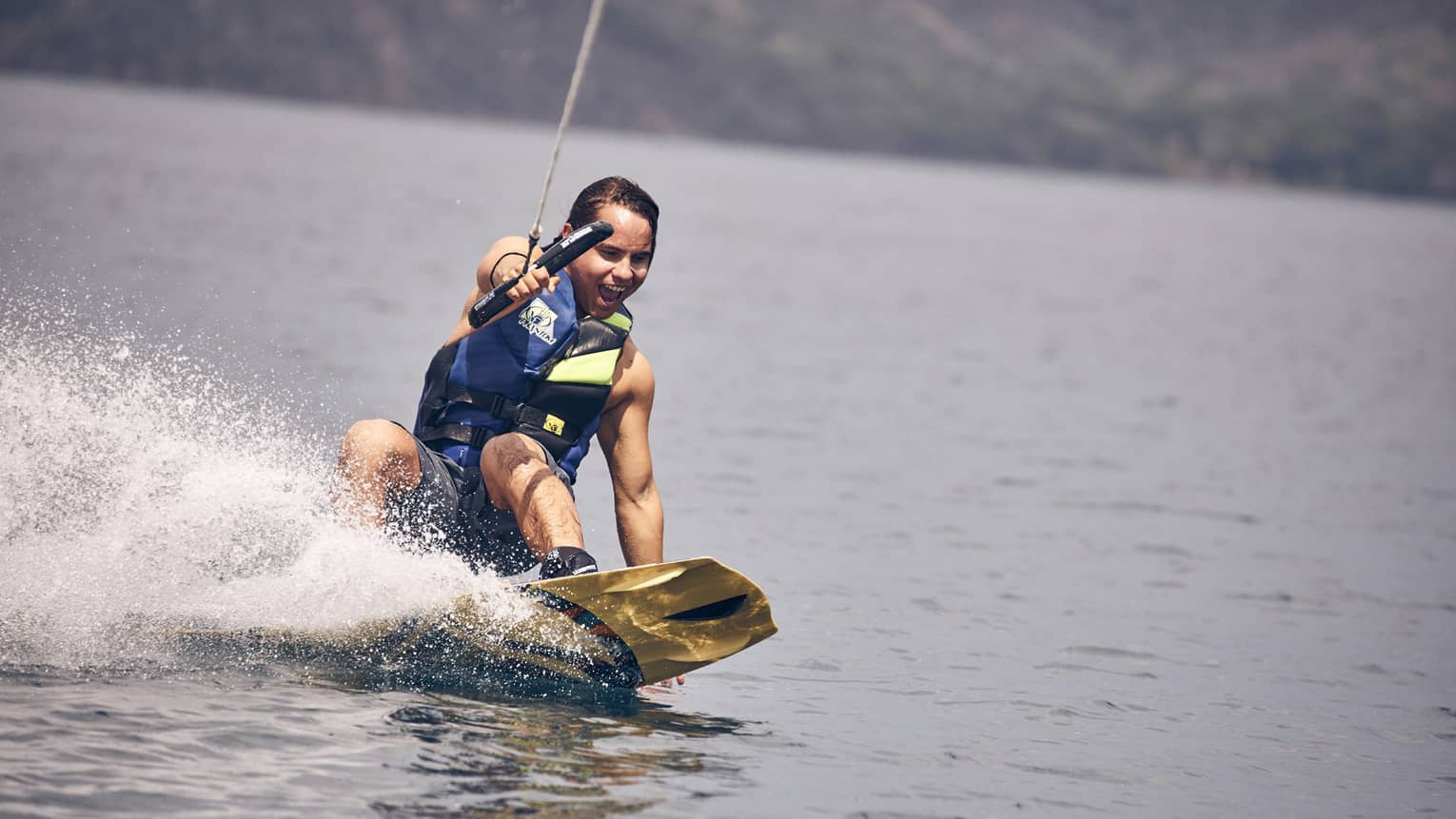 Young man wearing a blue and green life vest and blue swim trunks wakeboarding through the water