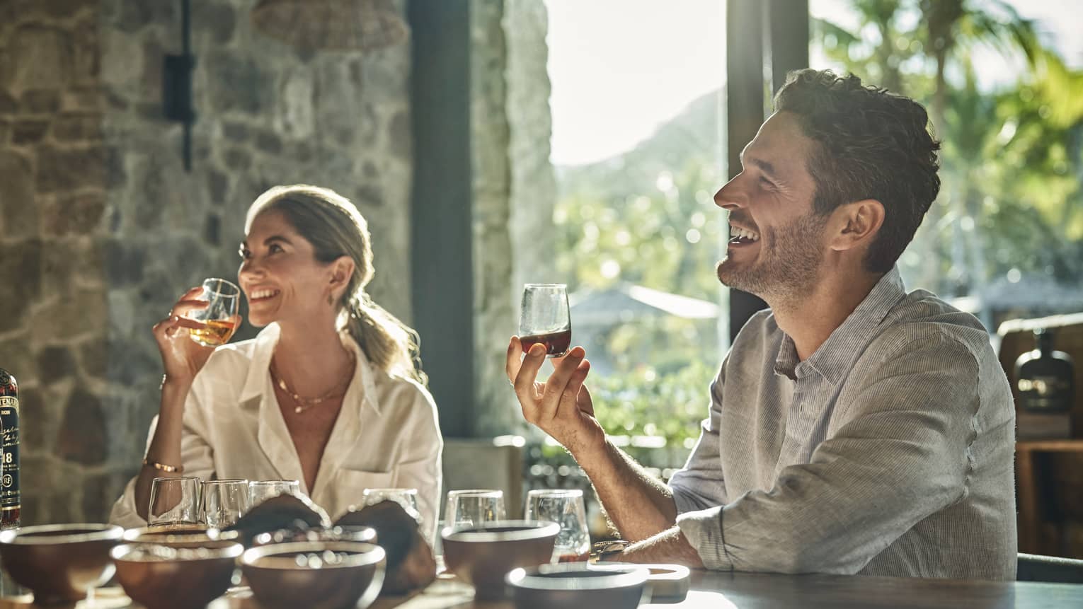 A man and a woman sit at a counter looking up and smiling and each holding a glass of rum with various other glasses and ingredients sit in front of them on the counter