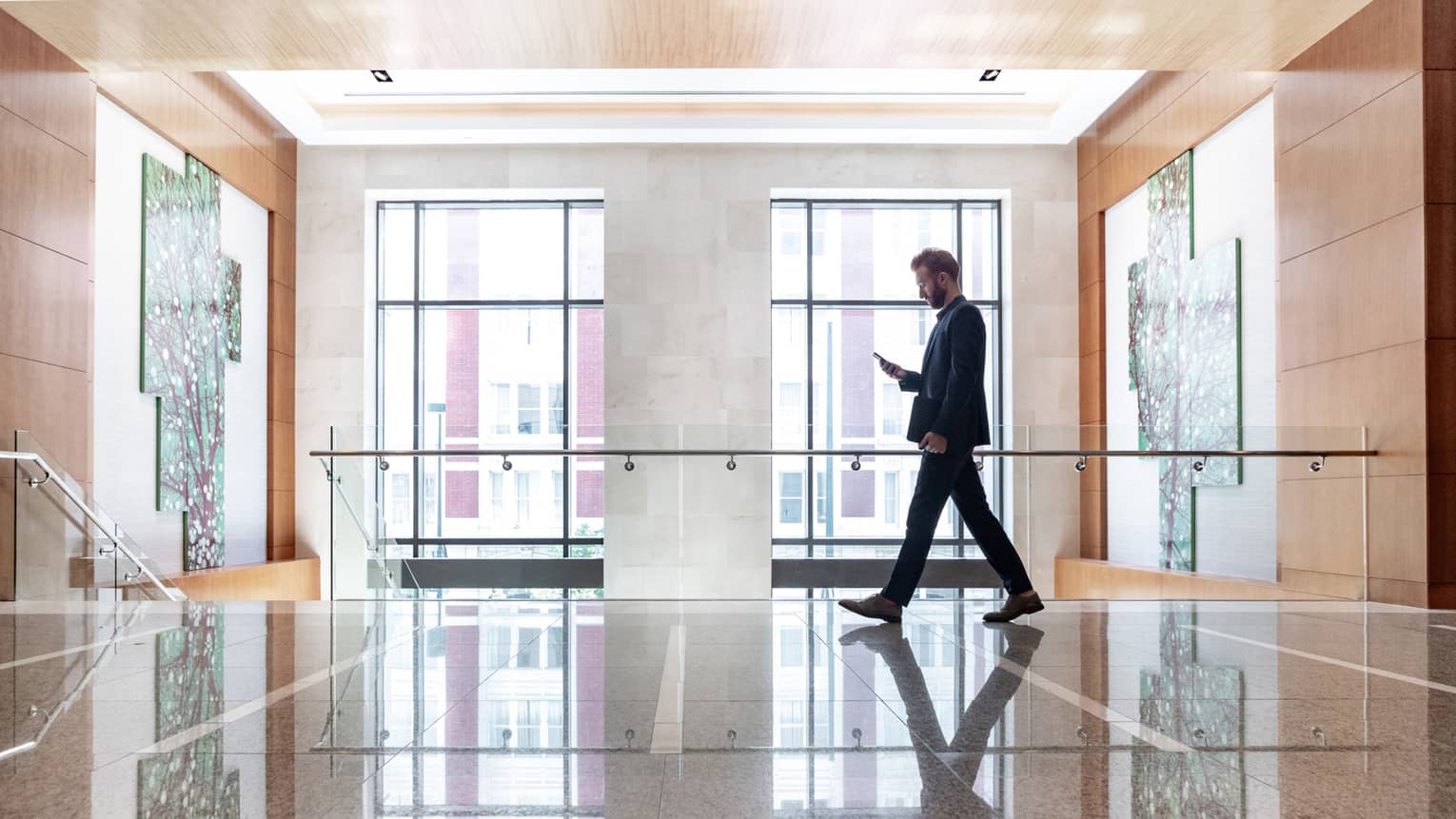 A man looking at his phone while he walks across a lobby area with large windows behind him
