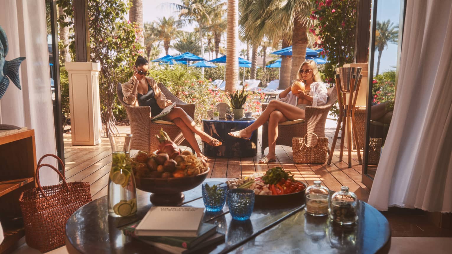 Two people sitting in rattan chairs with round table in forefront and blue pool umbrellas in backdrop