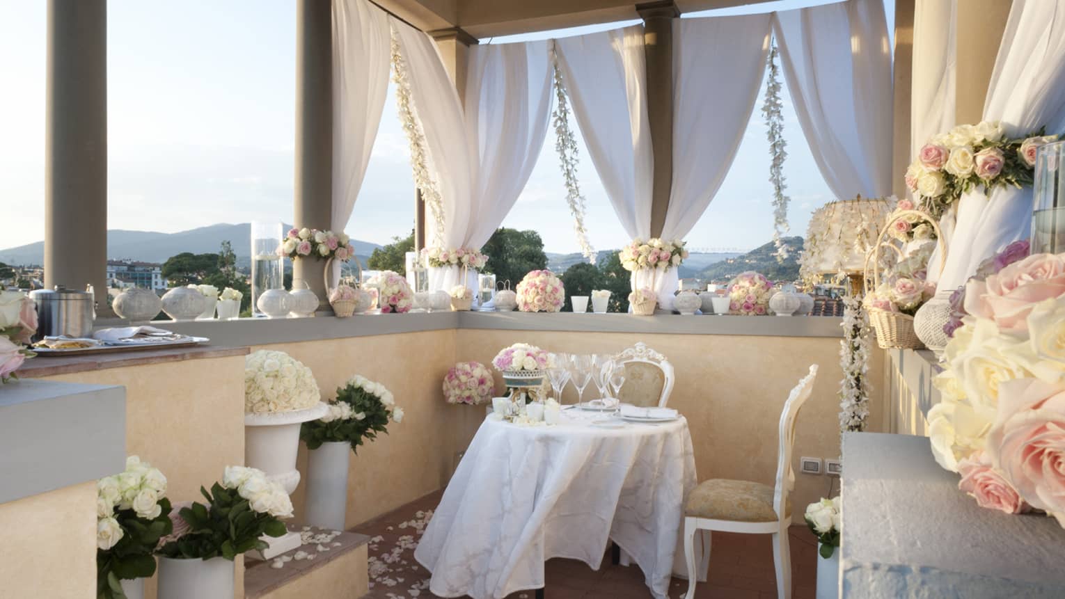 A romantic table set for two on a small terrace lined with pale roses and candles and a view of the city skyline beyond.