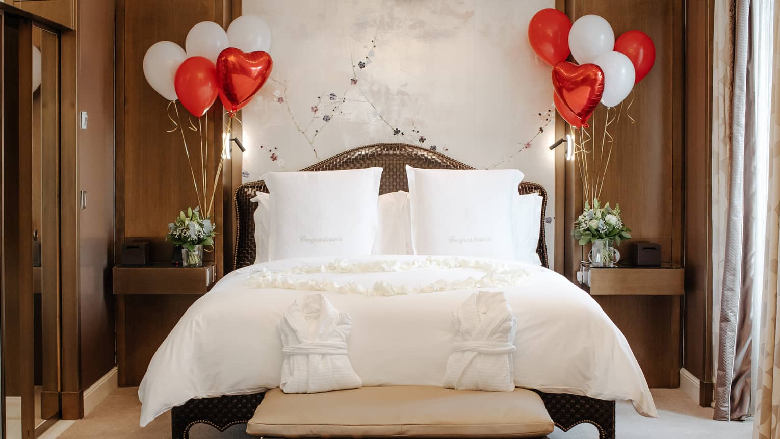 White Four Seasons bed against white wall, flanked with red and white balloons, and two white robes on foot bench
