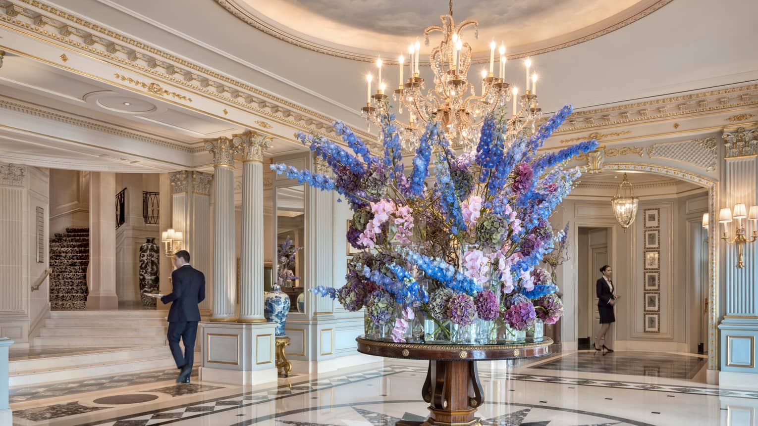 Lobby with marble art deco floor, white pillars with gold trim, round table with large, elaborate floral display
