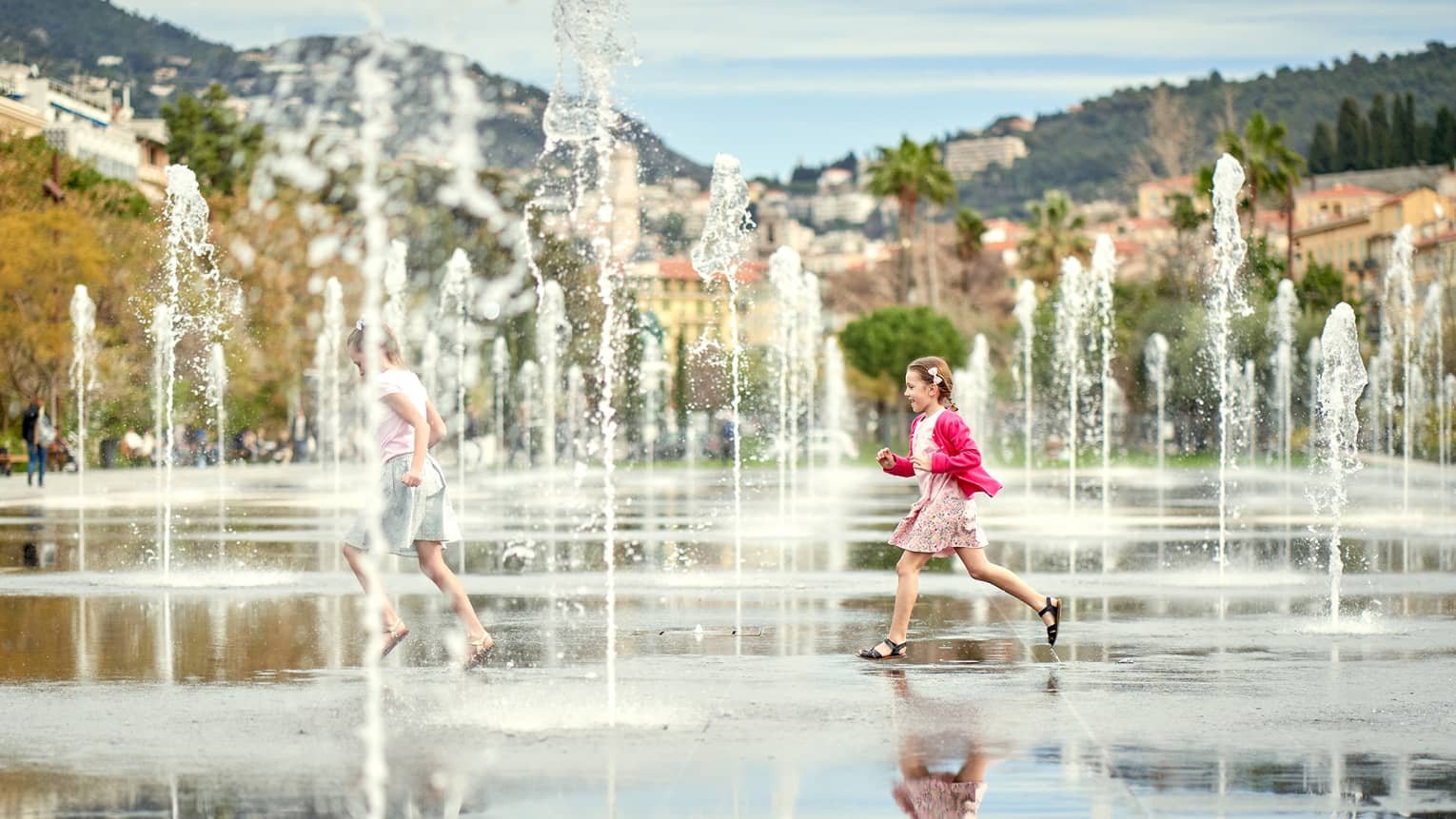 Two children running through water spouts, Riviera hillside in backdrop