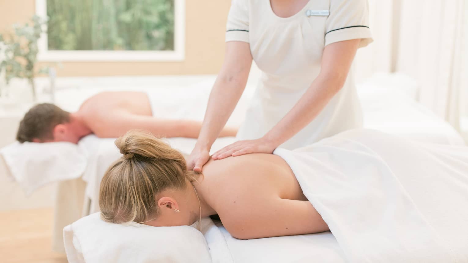 Couple laying on massage tables, spa staff massages woman's shoulders 