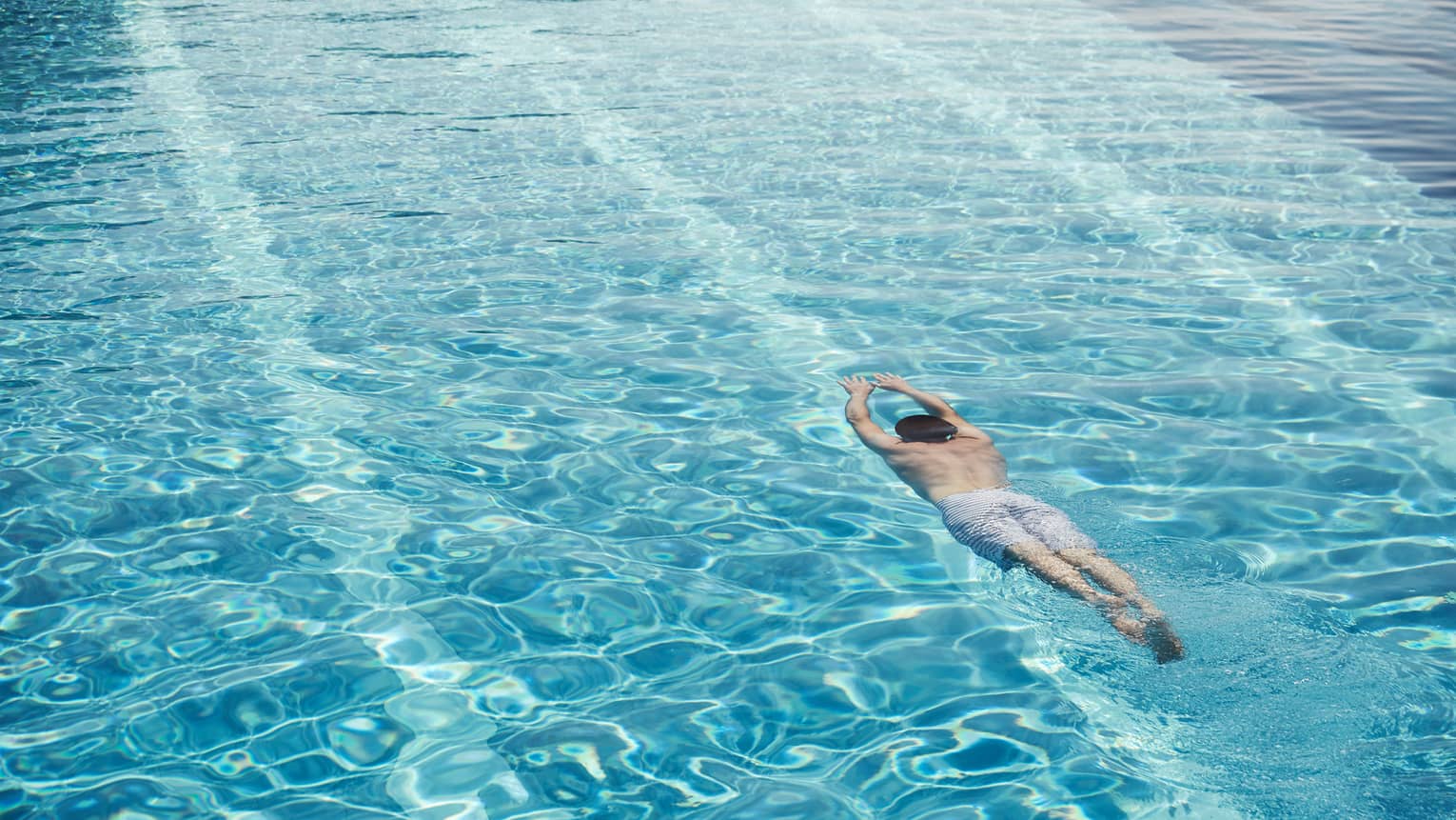 Man in swim shorts with arms outstretched dives underwater in blue swimming pool