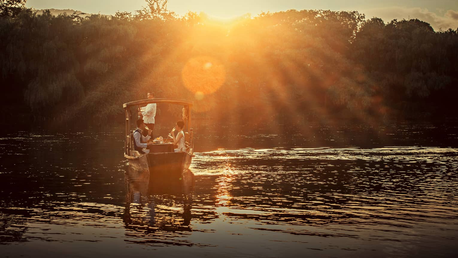 View from behind of a rustic wooden boat on a calm pond, under a golden glow; a couple enjoys champagne at a table inside.