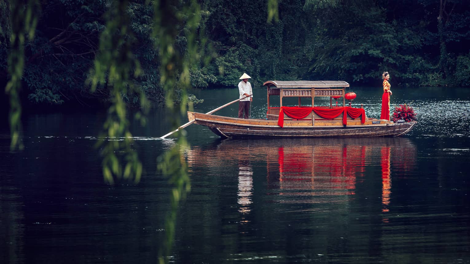 View across pond to bride wearing traditional red wedding gown at end of small boat