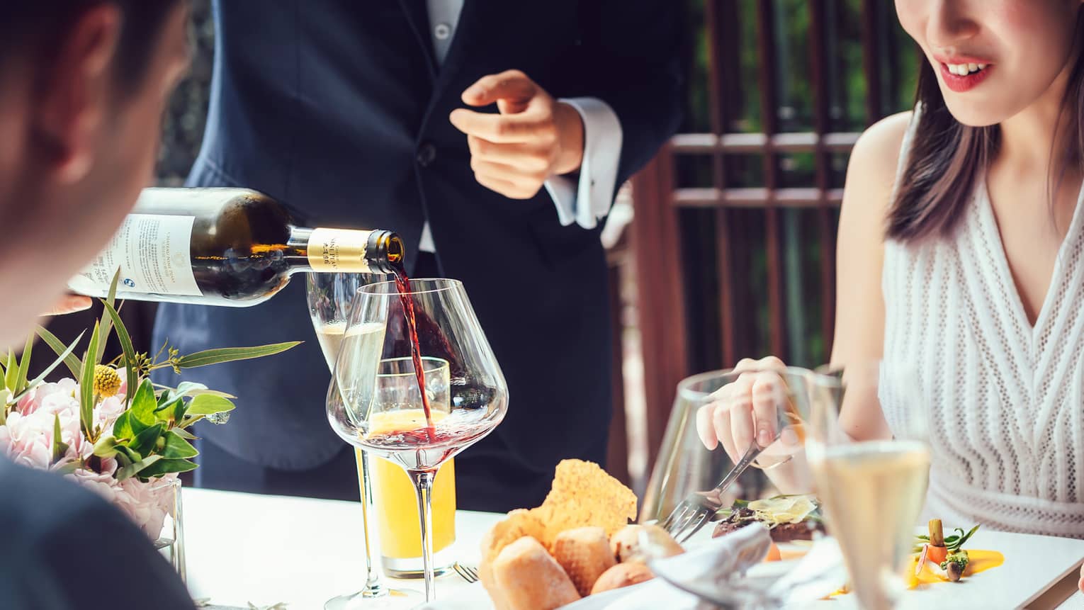 Server pouring red wine into large glass as couple dines at table