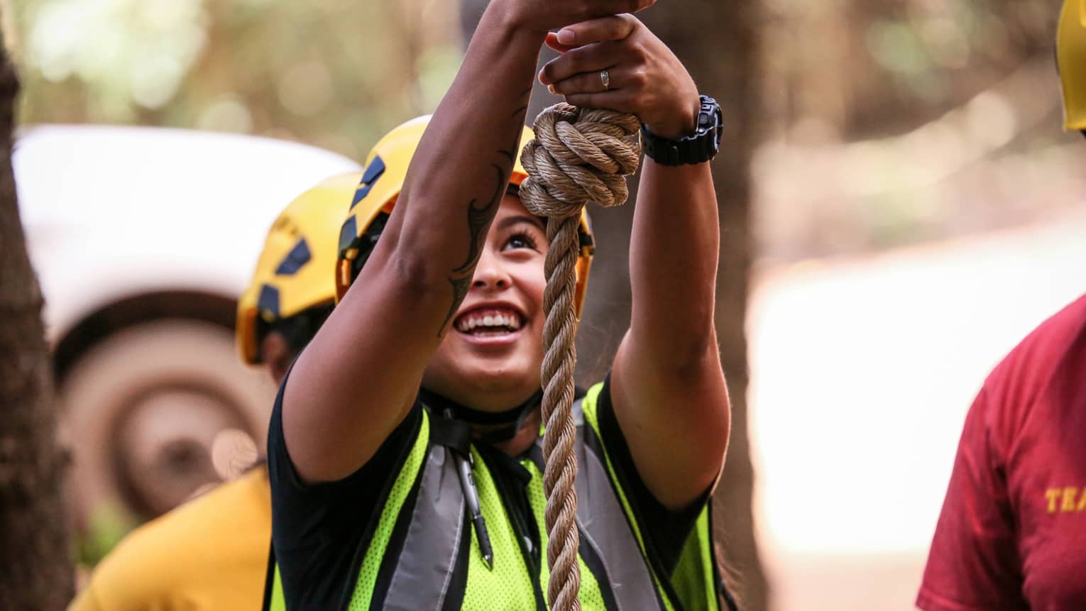 A woman in a helmet and safety vest smiles as she holds on to a rope