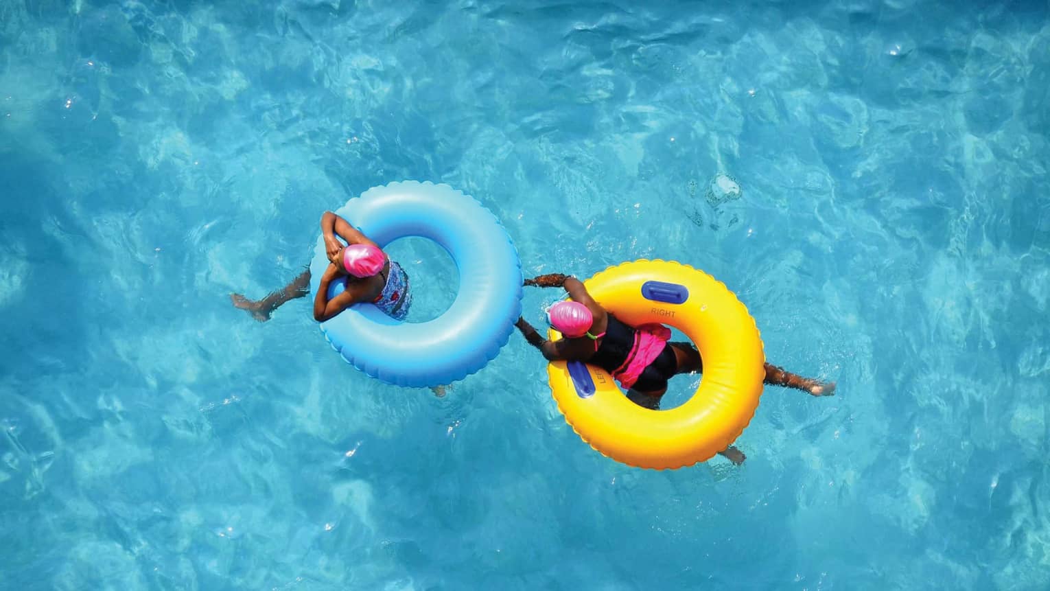 Aerial view of two swimmers in pink caps floating in blue and yellow inner tubes