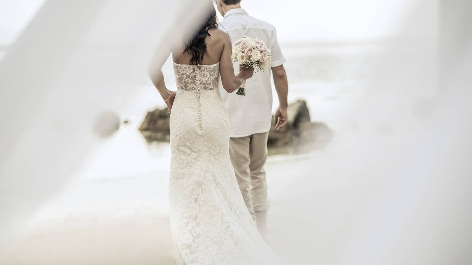 Bride wearing wedding dress and groom wearing white shirt walk on beach