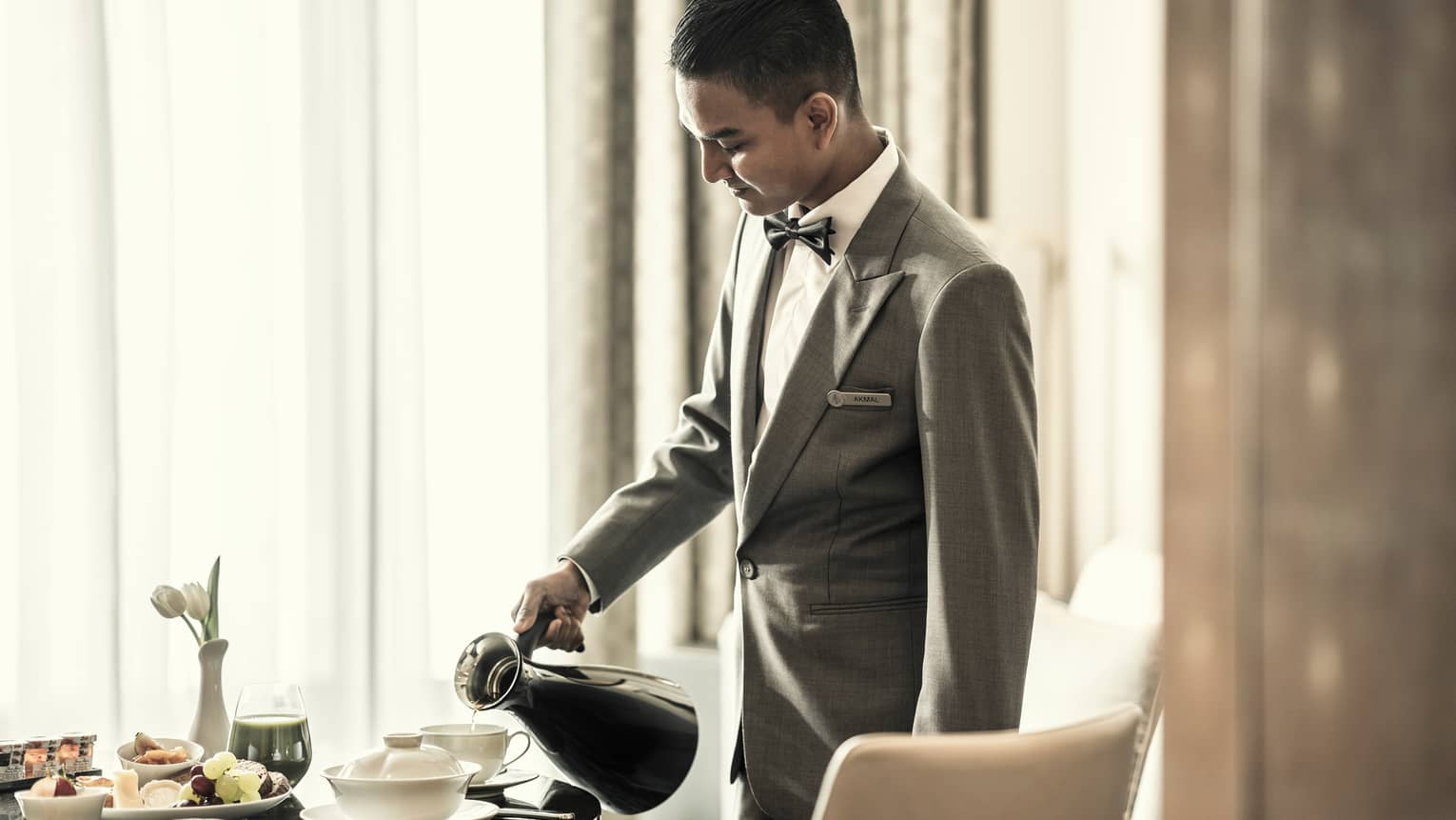 Hotel staff wearing suit jacket and bow tie pours coffee at in-room dining table