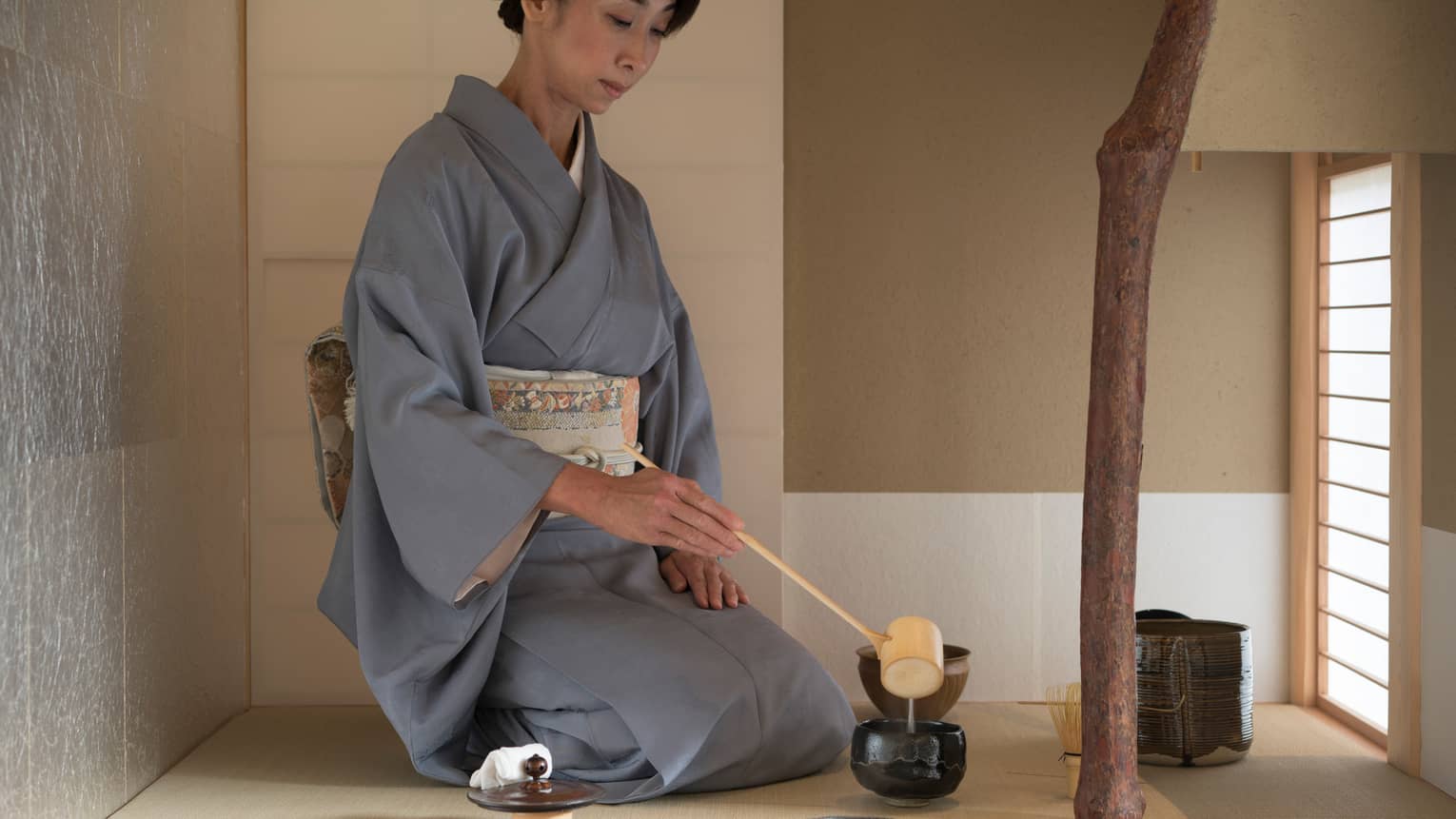 Woman in robe kneels on floor, ladles tea into cup with wood spoon