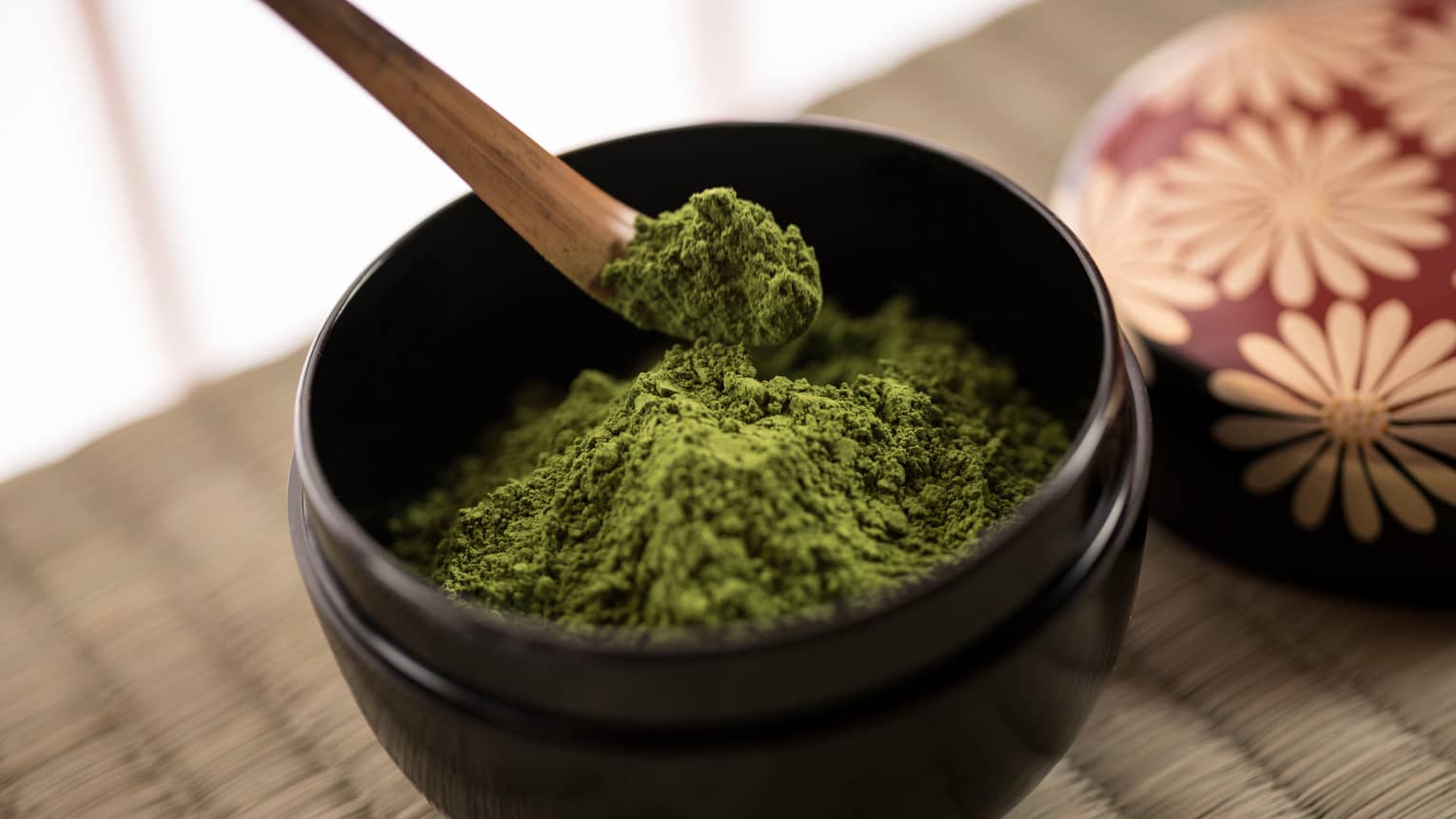 Close-up of green matcha tea powder in small black bowl, wood spoon