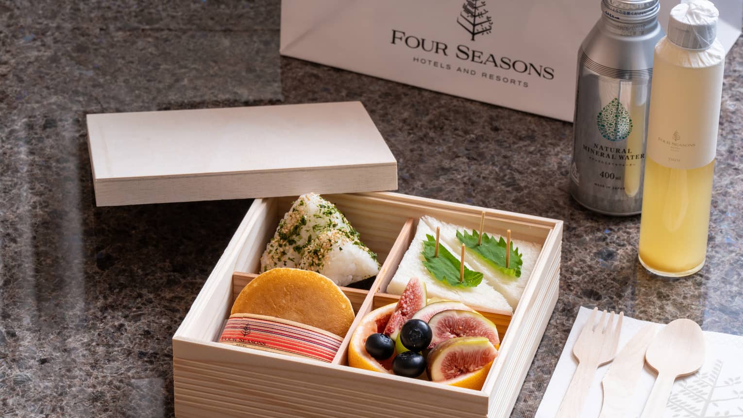 A wooden Four Seasons bento box with four compartments holding sandwiches, fruits and delicacies, beverage bottles nearby.