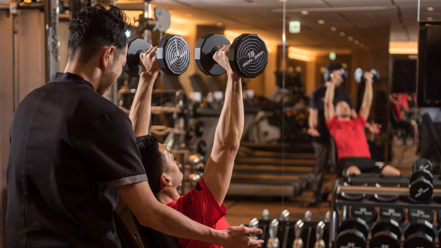 Man in chair in front of mirror lifts two 15 lb. weights above his head while personal trainer assists