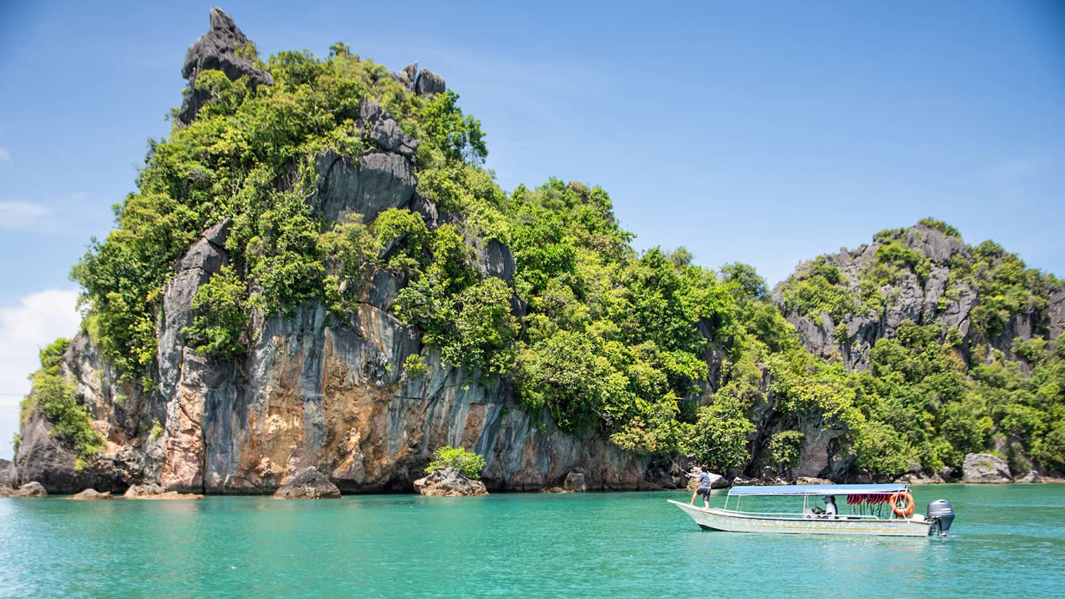 Guest fishing on a boat in turquoise water floating past a forested island with a tall craggy rock face under a blue sky.