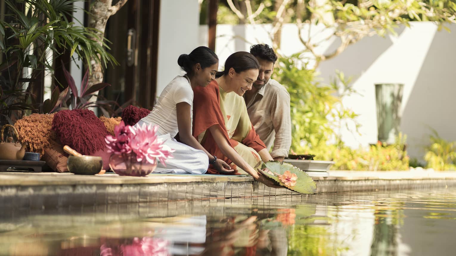 Two women and a man kneeling at a pond and releasing a floating lotus