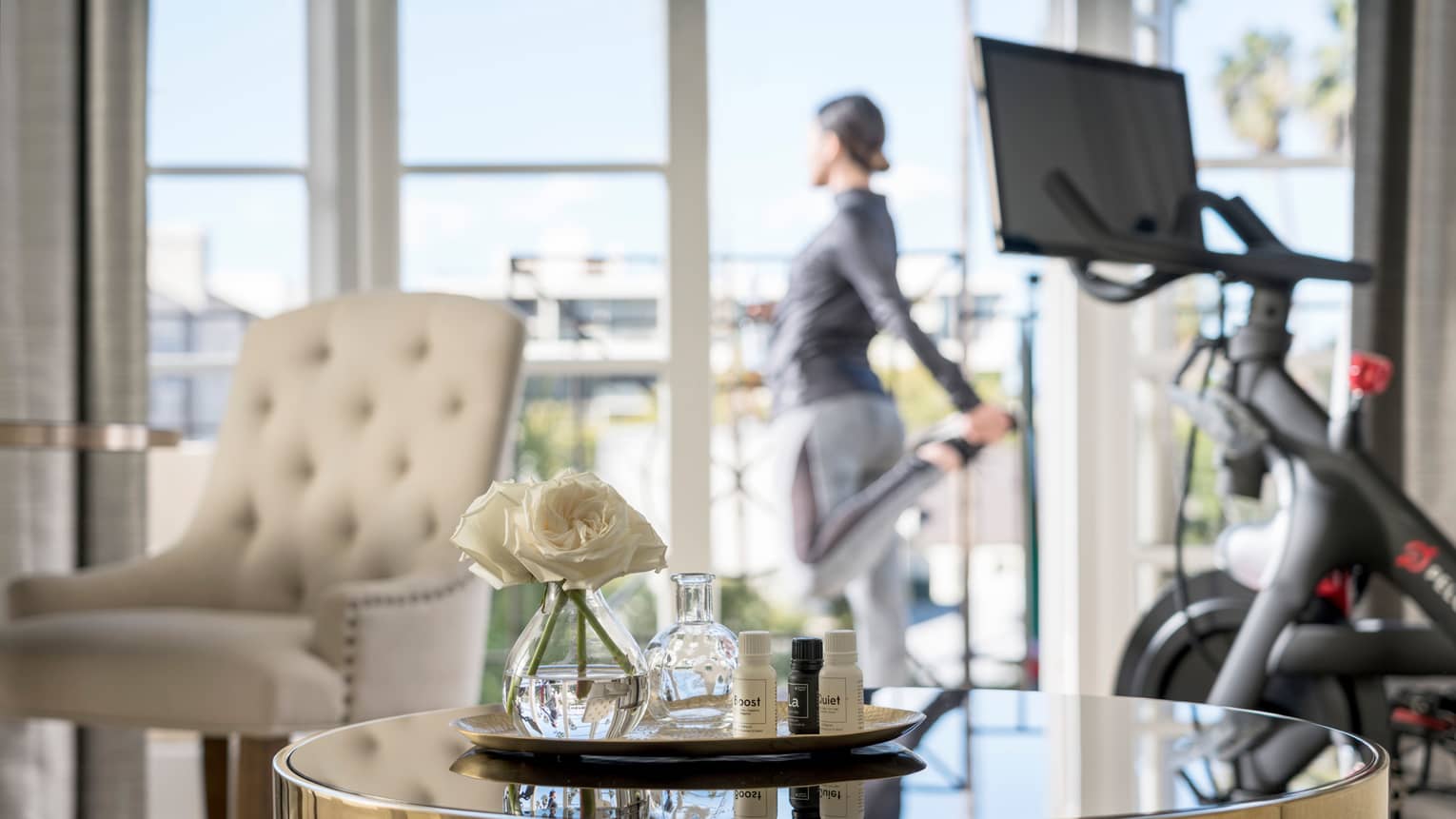 Spa tray with fresh white flowers on table by treadmill, woman stretching at balcony door