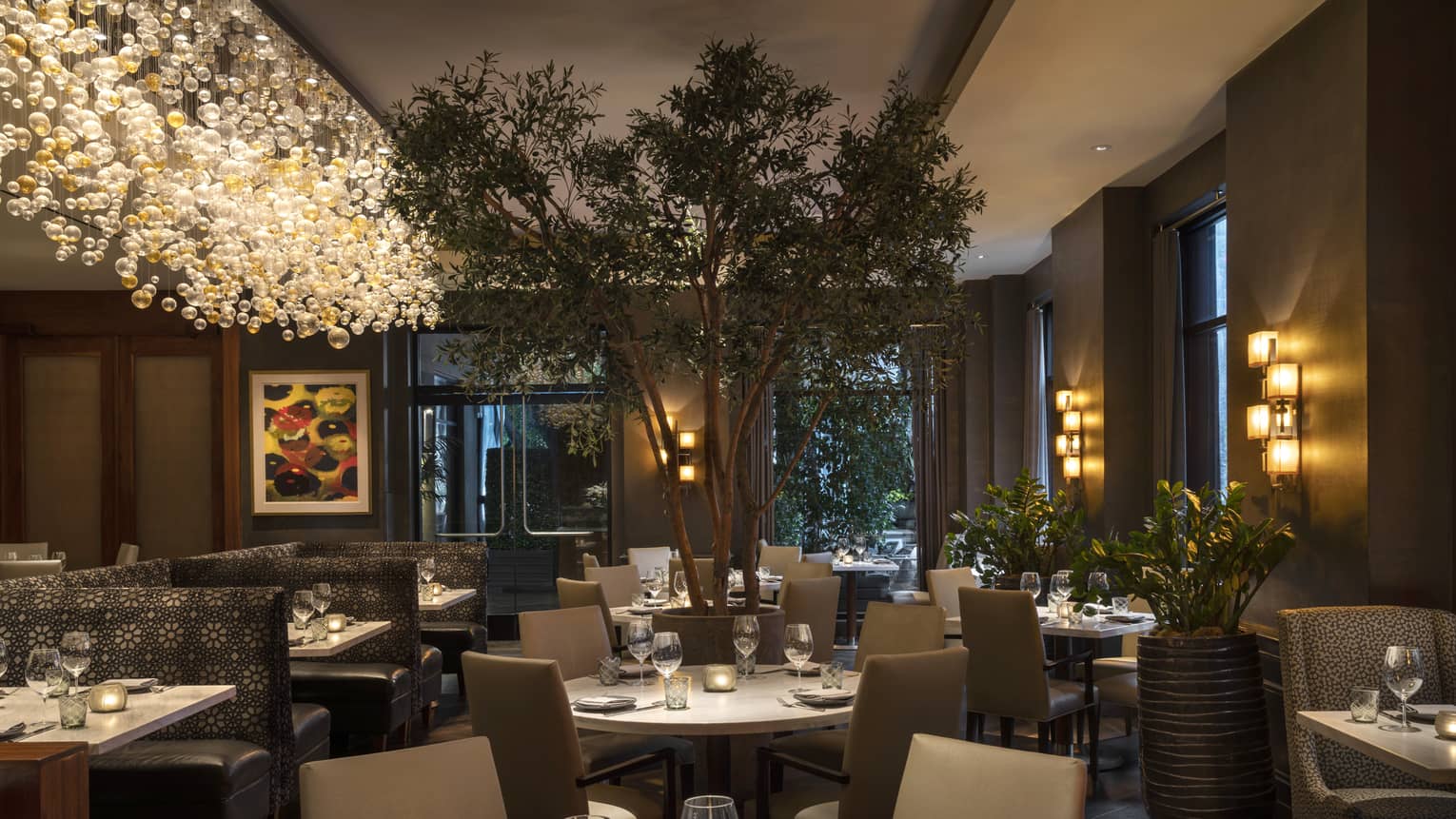 Indoor restaurant in the evening with square tables, taupe chairs, large tree, long hanging chandelier