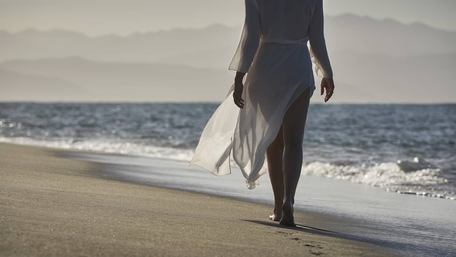 A woman walking down the shore of a beach next to waves running up on the shore.