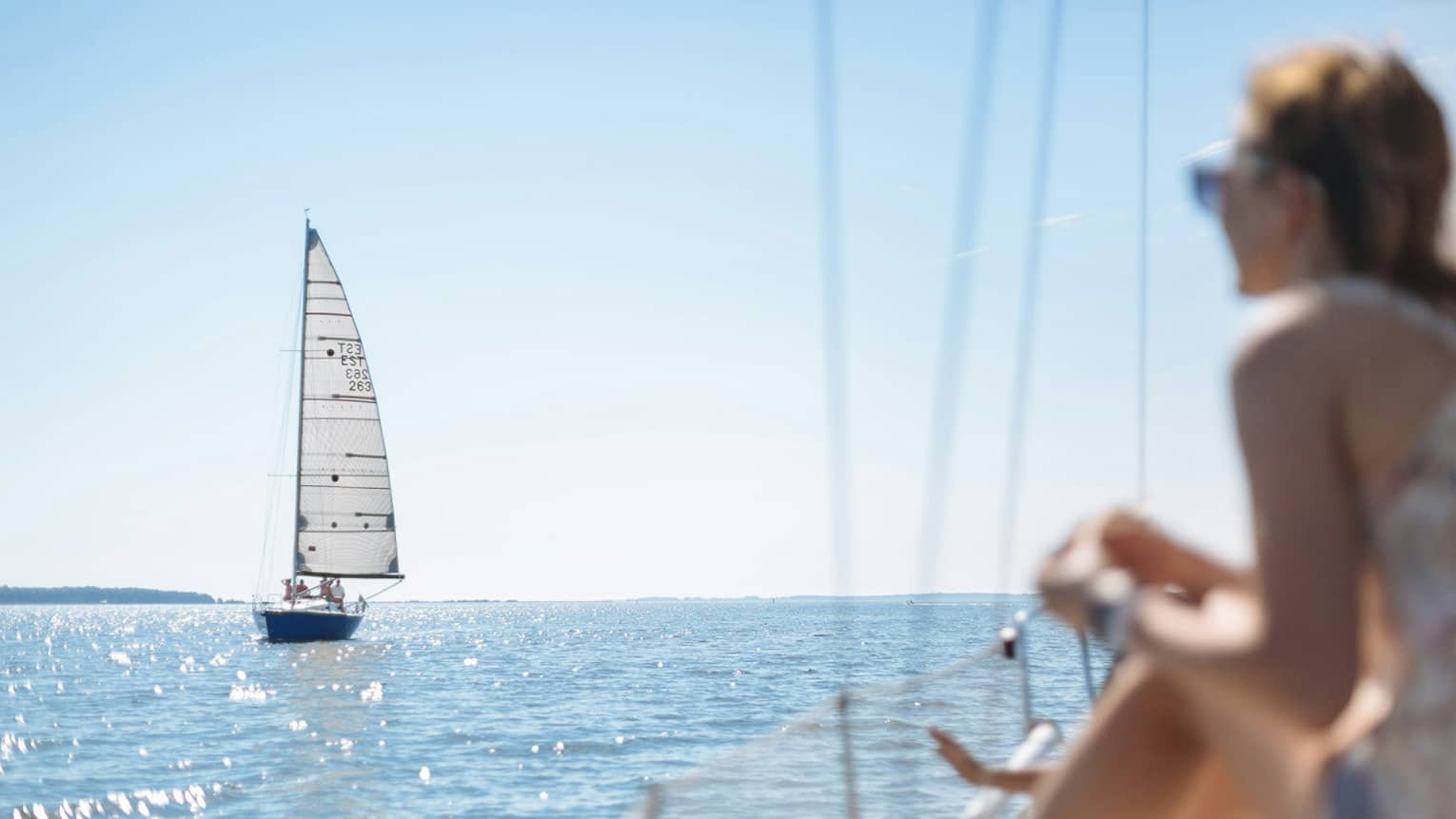 In the foreground, a passenger on the deck of a boat watches a distant sailboat over sparkling water in the bright sun.