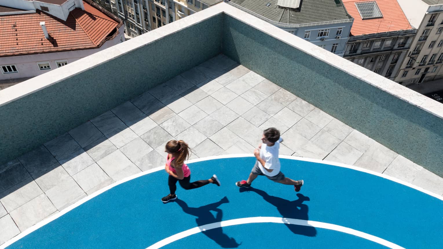 Aerial view of man and woman running on blue jogging track, city rooftops seen below