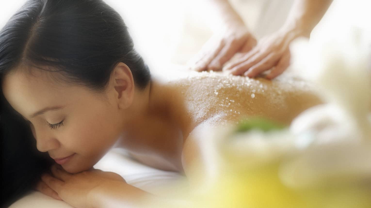 Woman lying down with eyes closed as hands massage salt scrub into shoulders
