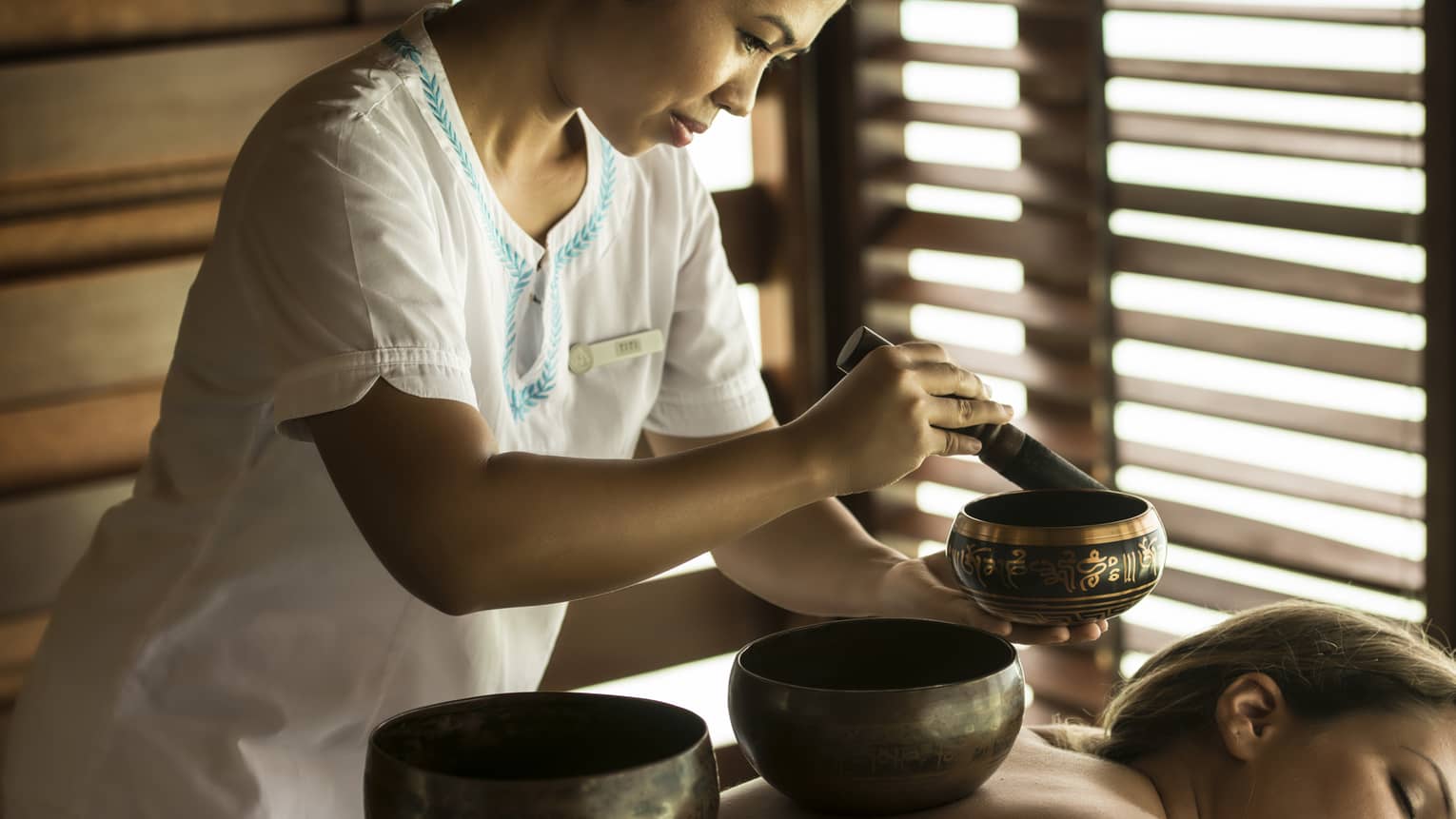 Guest with singing bowls on bare back lies on treatment table which sound specialist plays another singing bowl during a sound healing session