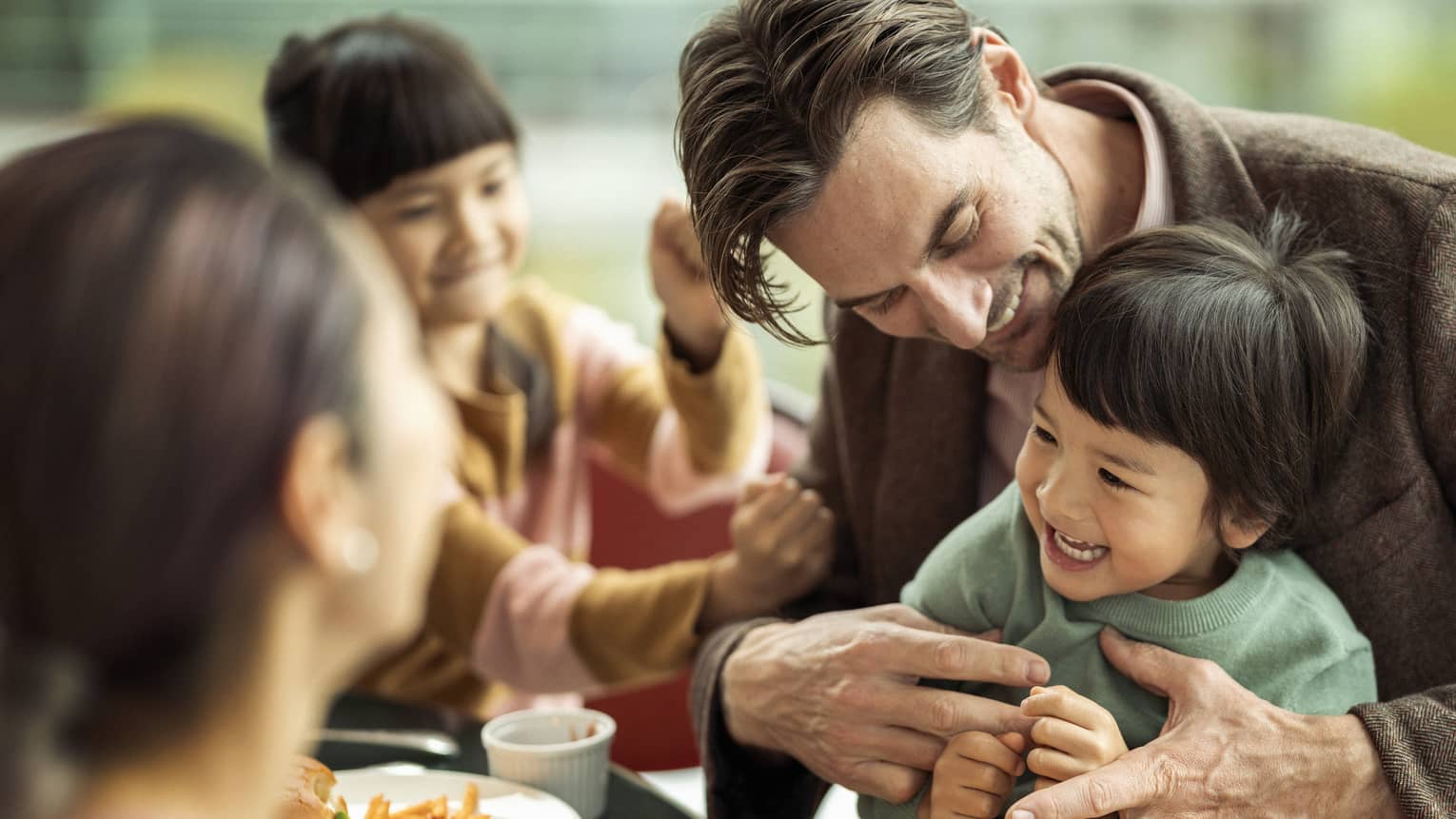 Smiling man holds giggling young boy on his lap at restaurant as woman and young girl look on