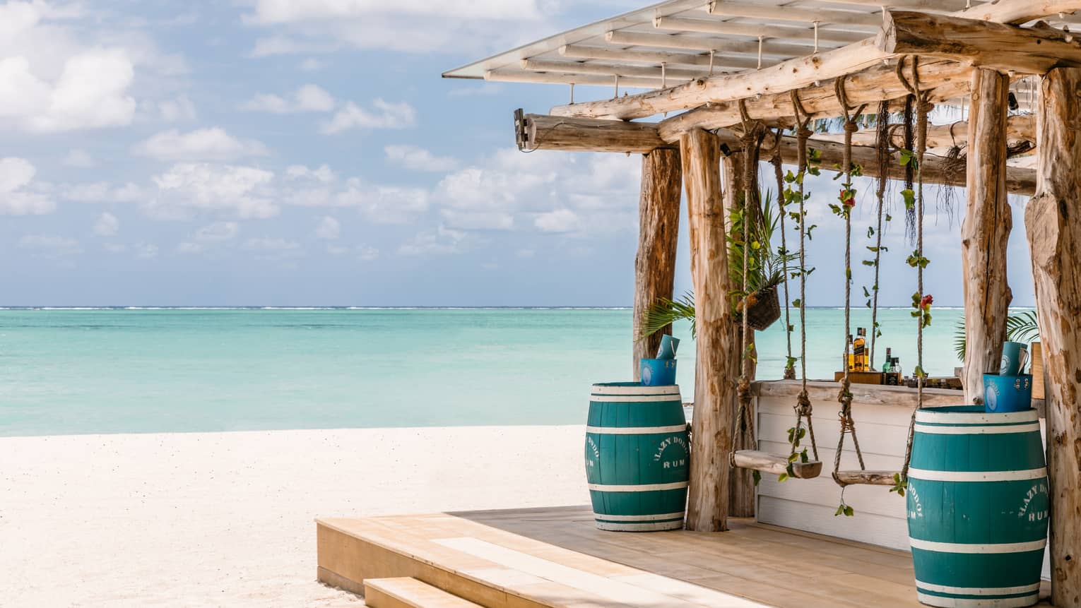 Wooden bar with pergola, two blue barrels, two swings on beach, aqua views, cloud dotted blue sky