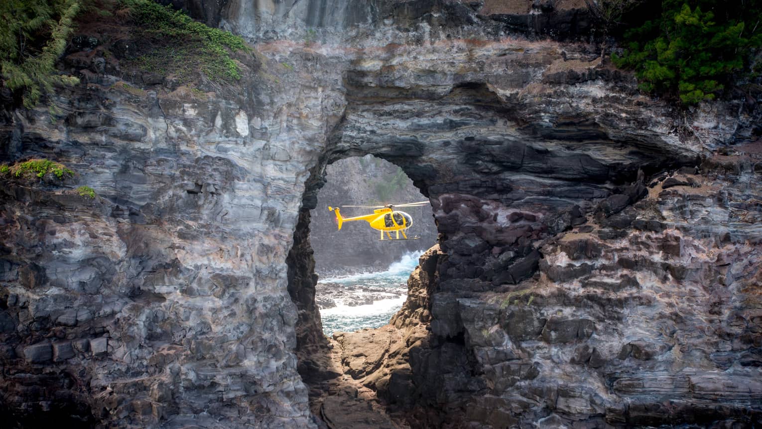 A sideview of a yellow holicopter, seen through an opening in a rock structure.