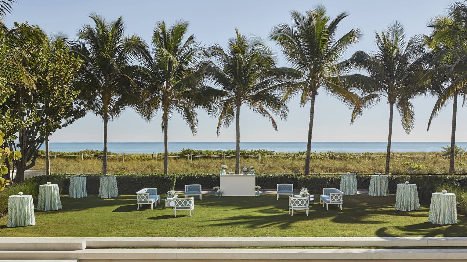 Blue outdoor sofas surround white bar, cocktail tables with long tablecloths on grass surrounded by tall palm trees with an ocean view