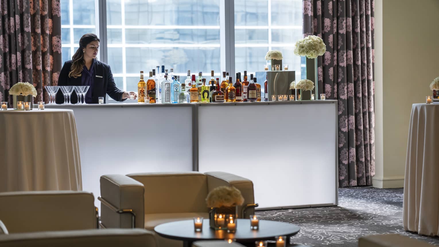 Bartender stands at liquor bottle-lined bar in front of window near seating area