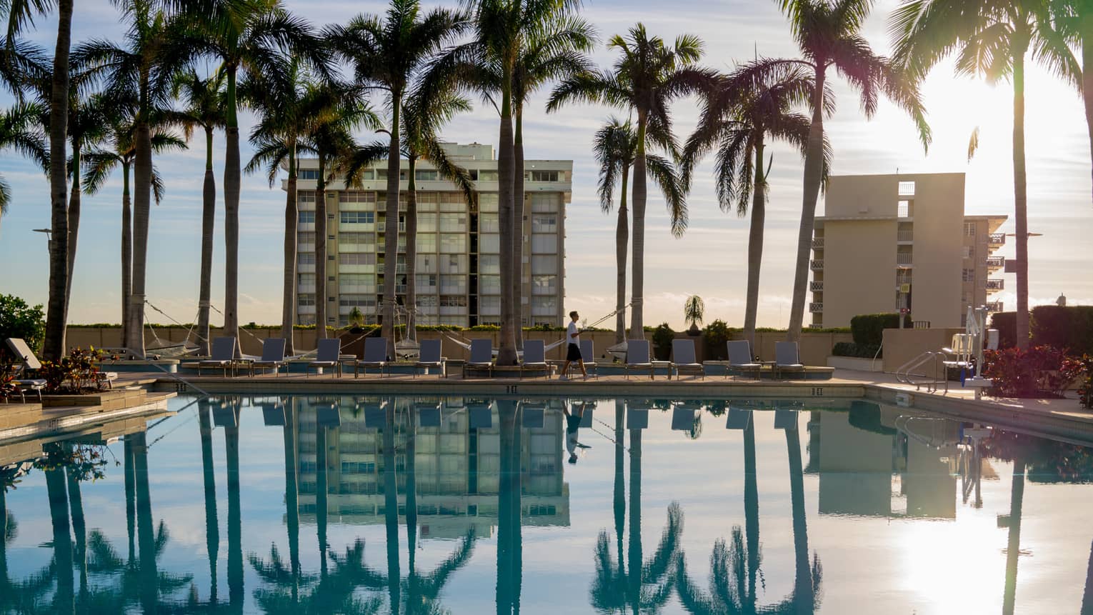 A staff member walking next to a pool on a sunny day with palm trees and building around it.