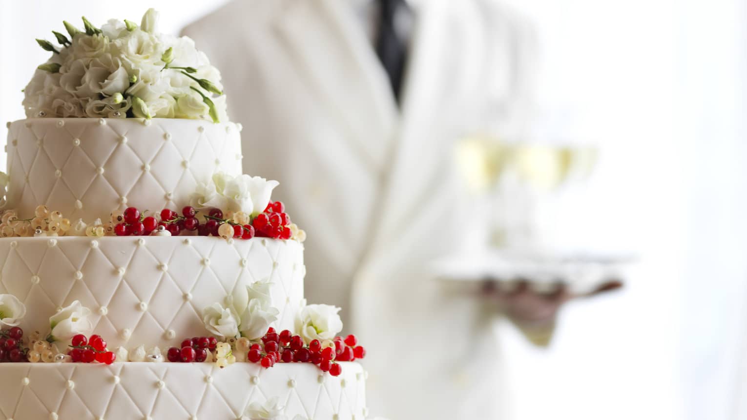 Waiter with tray of Champagne glasses behind white tiered wedding cake with flowers