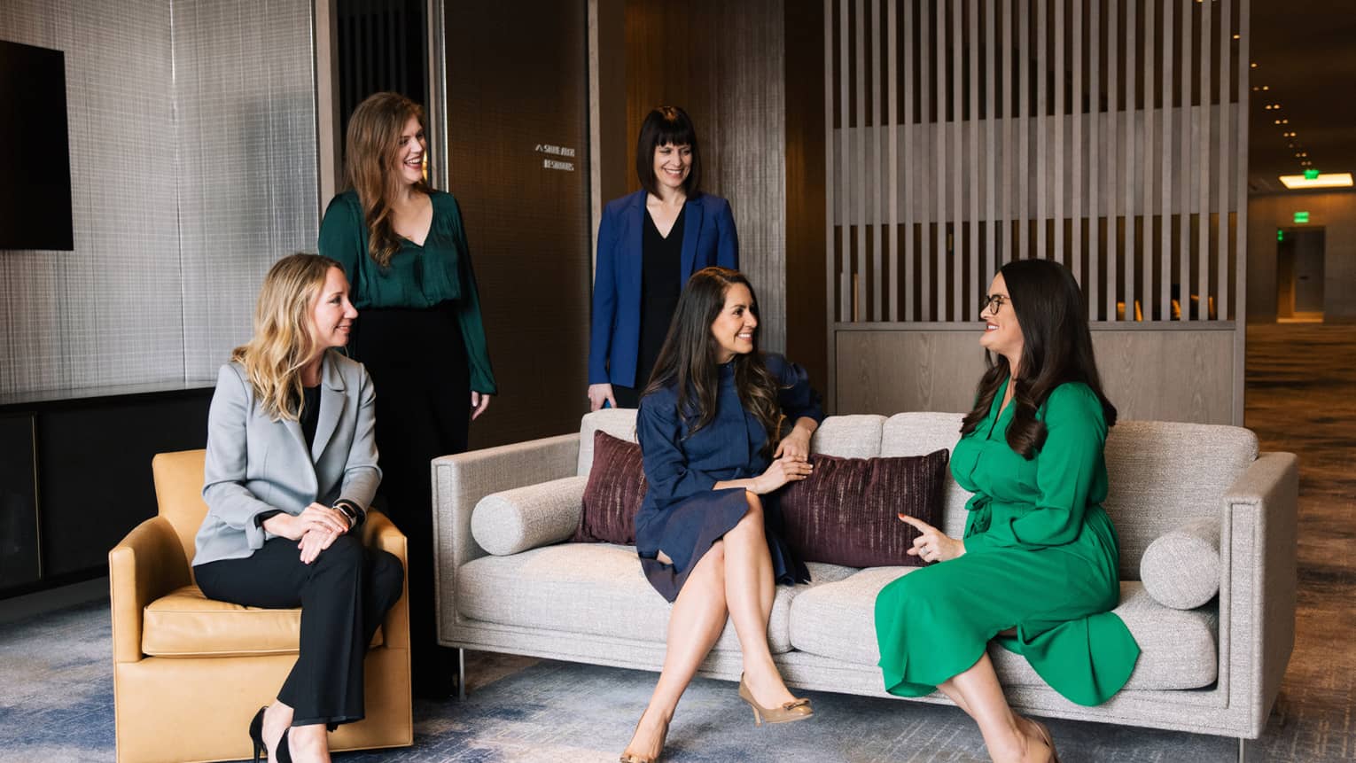 Two women sit on a sofa next to another woman in an armchair with two other women standing behind them