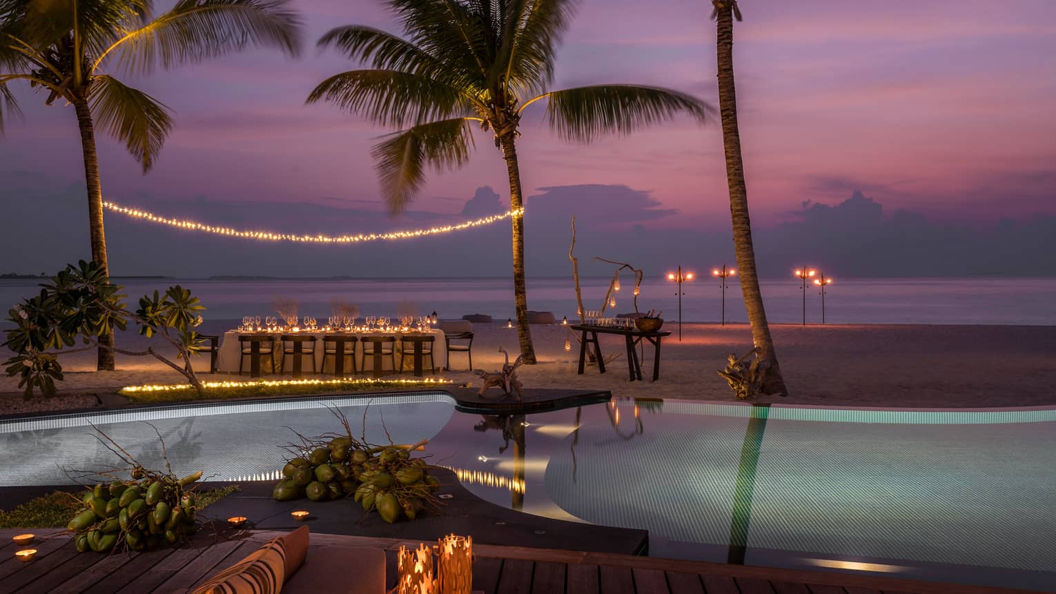 String of lights between palm tree over private dining table on beach by swimming pool at sunset
