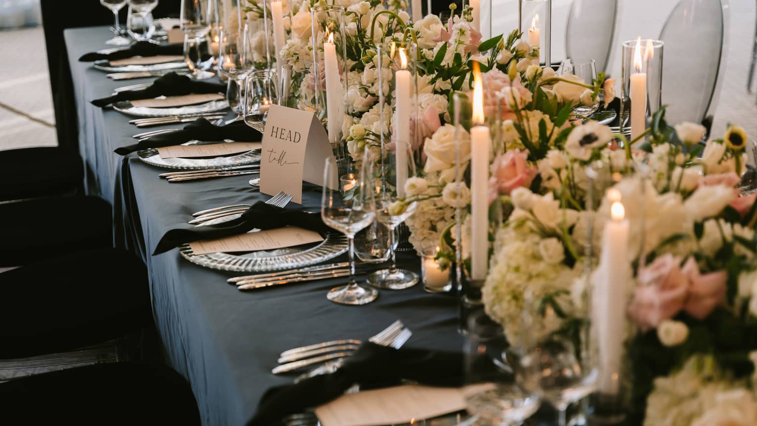 Long banquet table set for a wedding reception with a black table cloth, black napkins, glass chargers, white tapered candles and a dense centrepiece of white, pink and green florals
