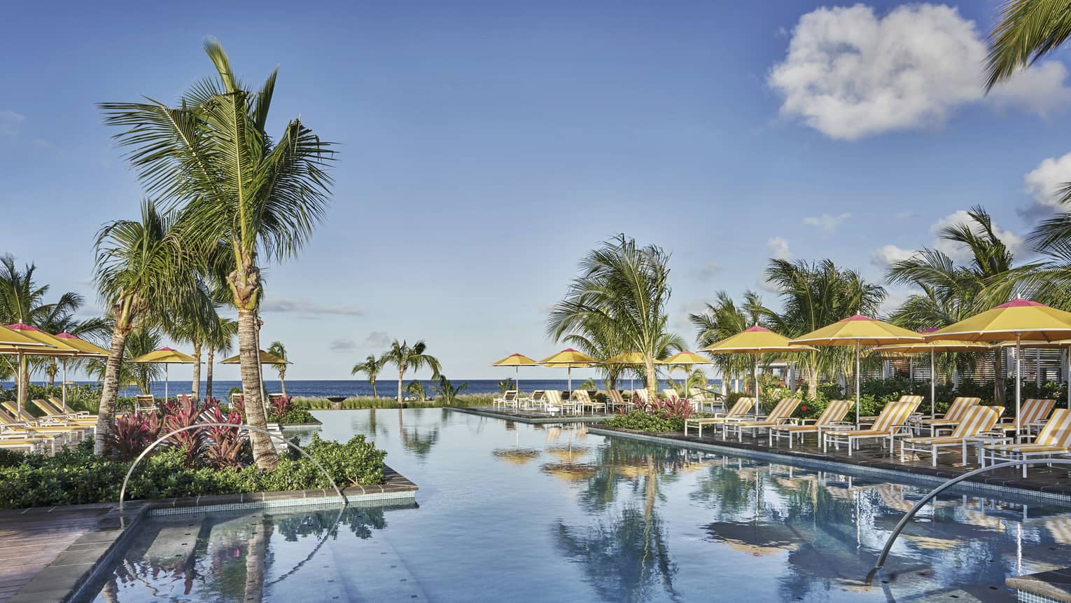 A calm pool reflecting the blue sky and palm trees, lined with lounge chairs and yellow umbrellas