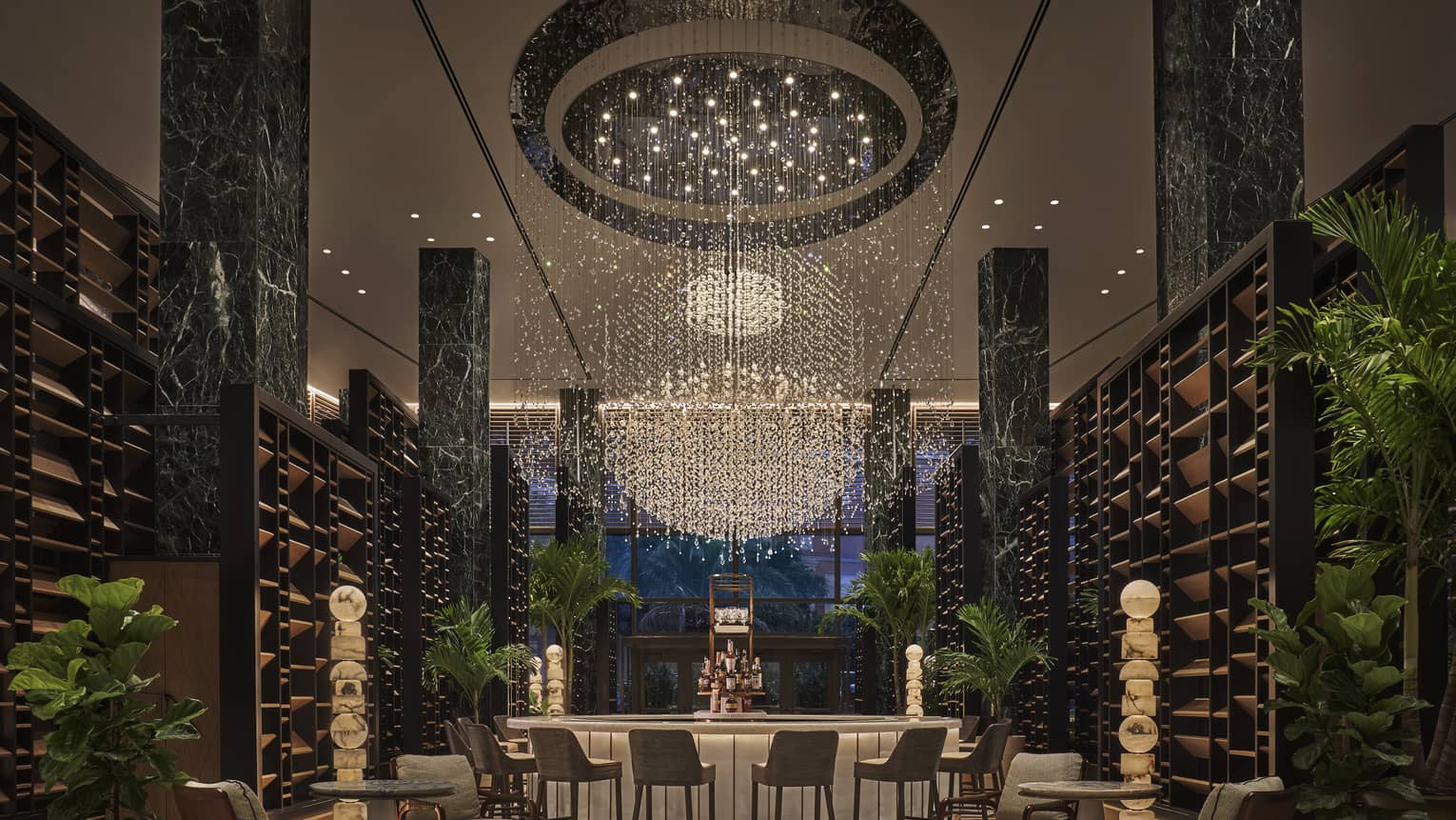 Chandelier Bar with six jade pillars flanked by wooden shelves and potted palms, glass-topped bar in the middle with six cocktail chairs, giant chandelier hanging overhead