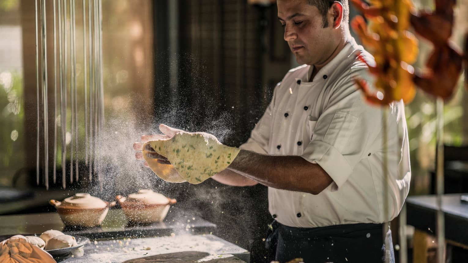 Chef at counter tosses dough with herbs, flour in air around him