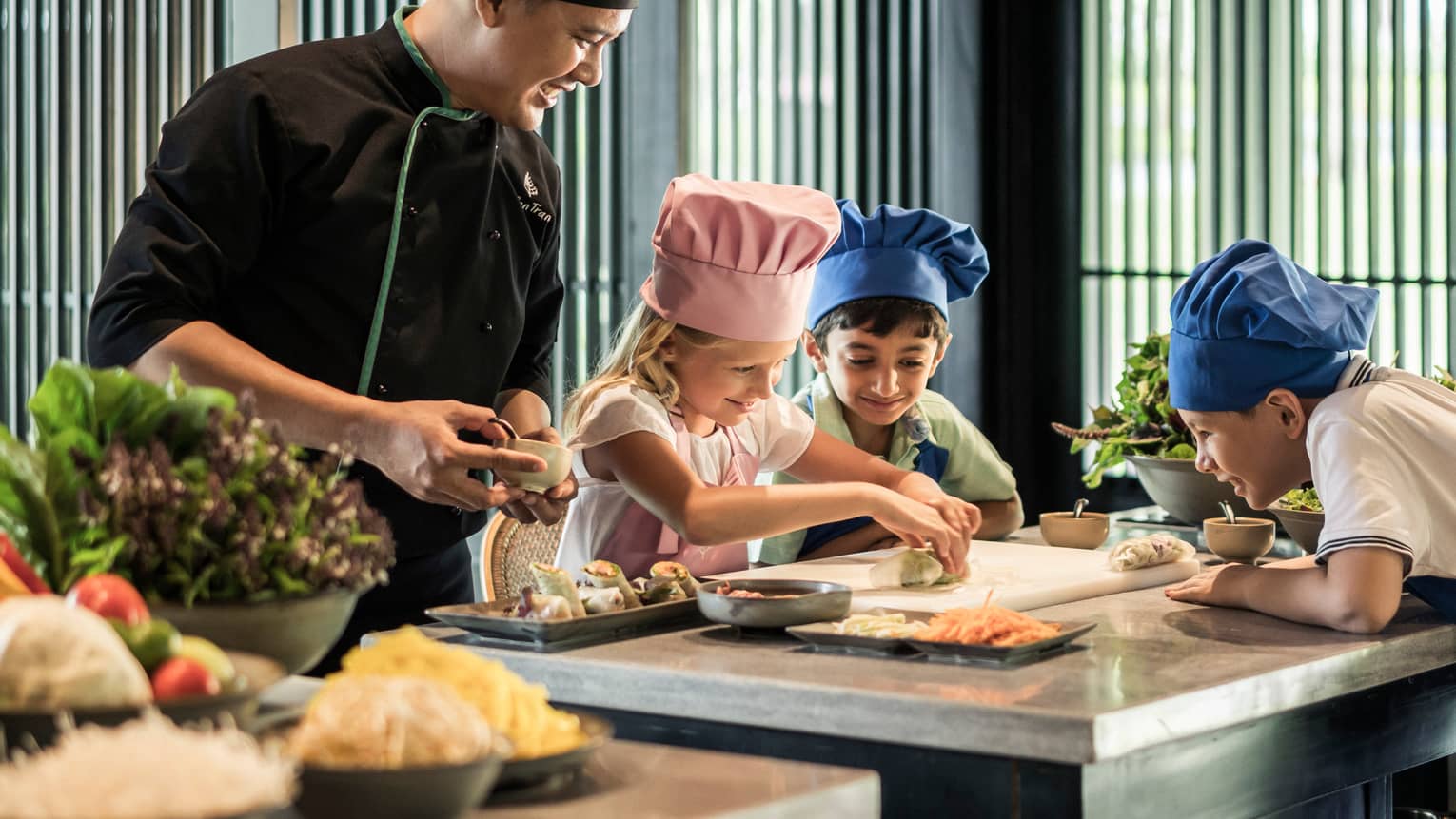 Chef watches as children wearing tall chef hats make spring rolls at counter