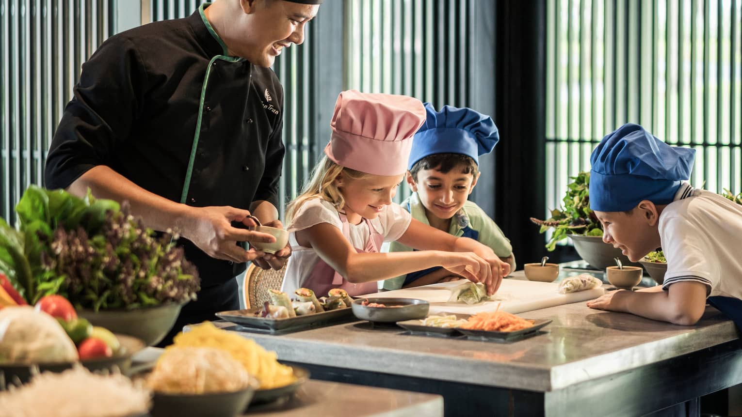 Chef watches as children wearing tall chef hats make spring rolls at counter
