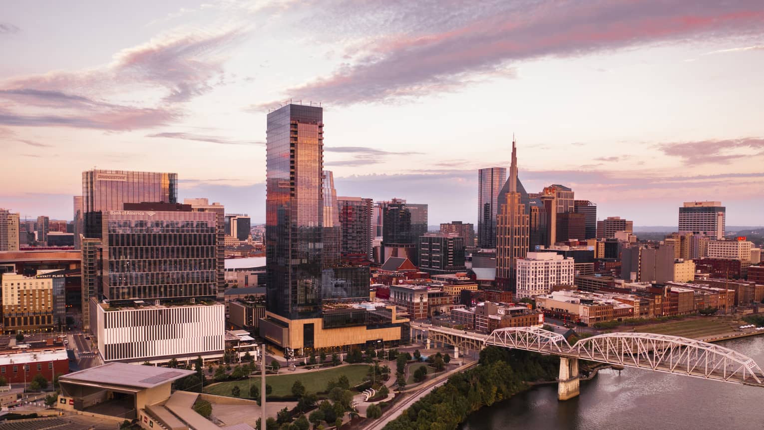 Aerial view of downtown Nashville, Tennessee at sunset, featuring modern skyscrapers, a river, a bridge, and a park with greenery in the foreground.