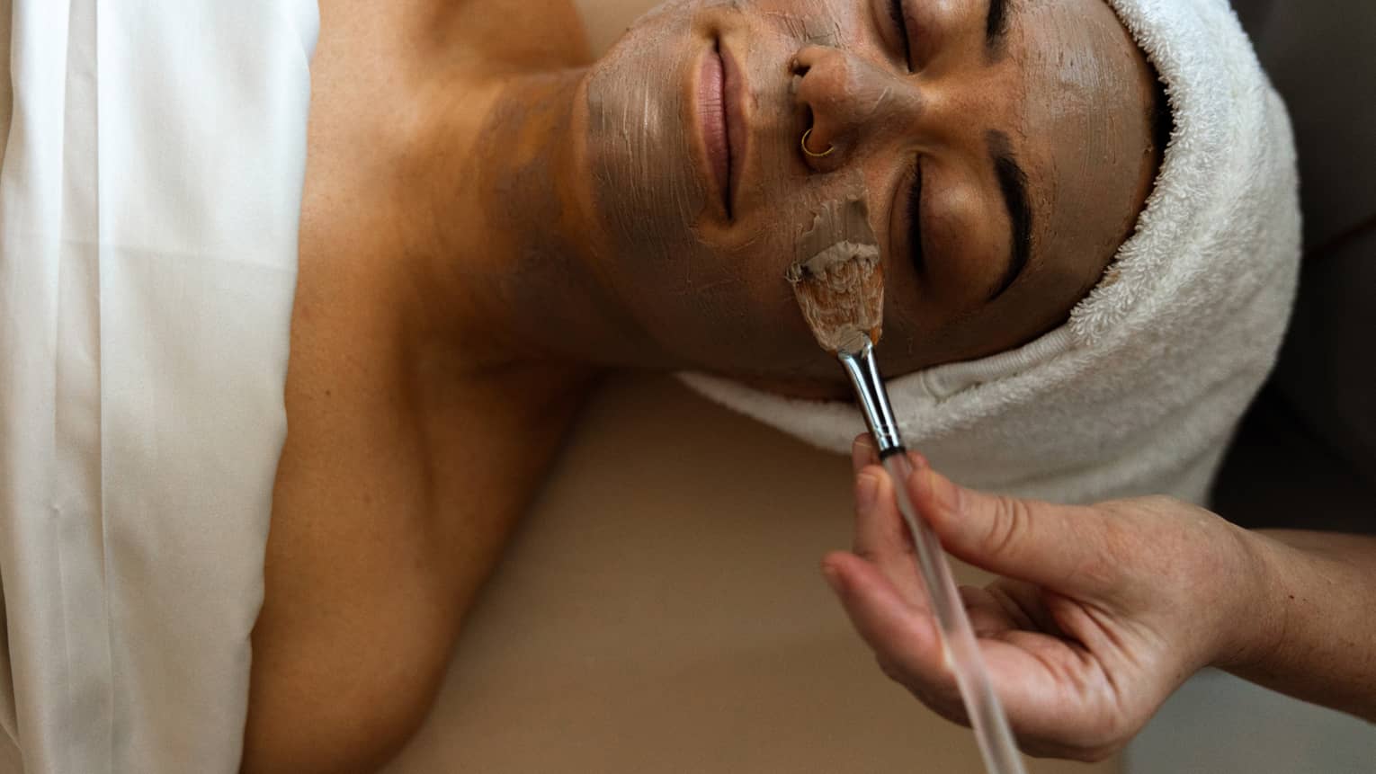 Woman with hair wrapped in towel getting a face masque put on.
