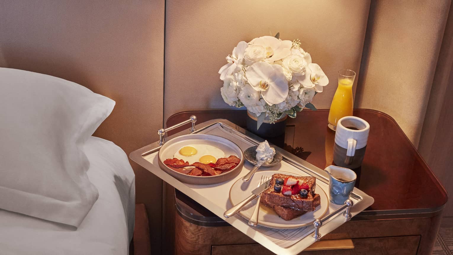 Tray on nightstand with eggs, bacon and french toast on it.