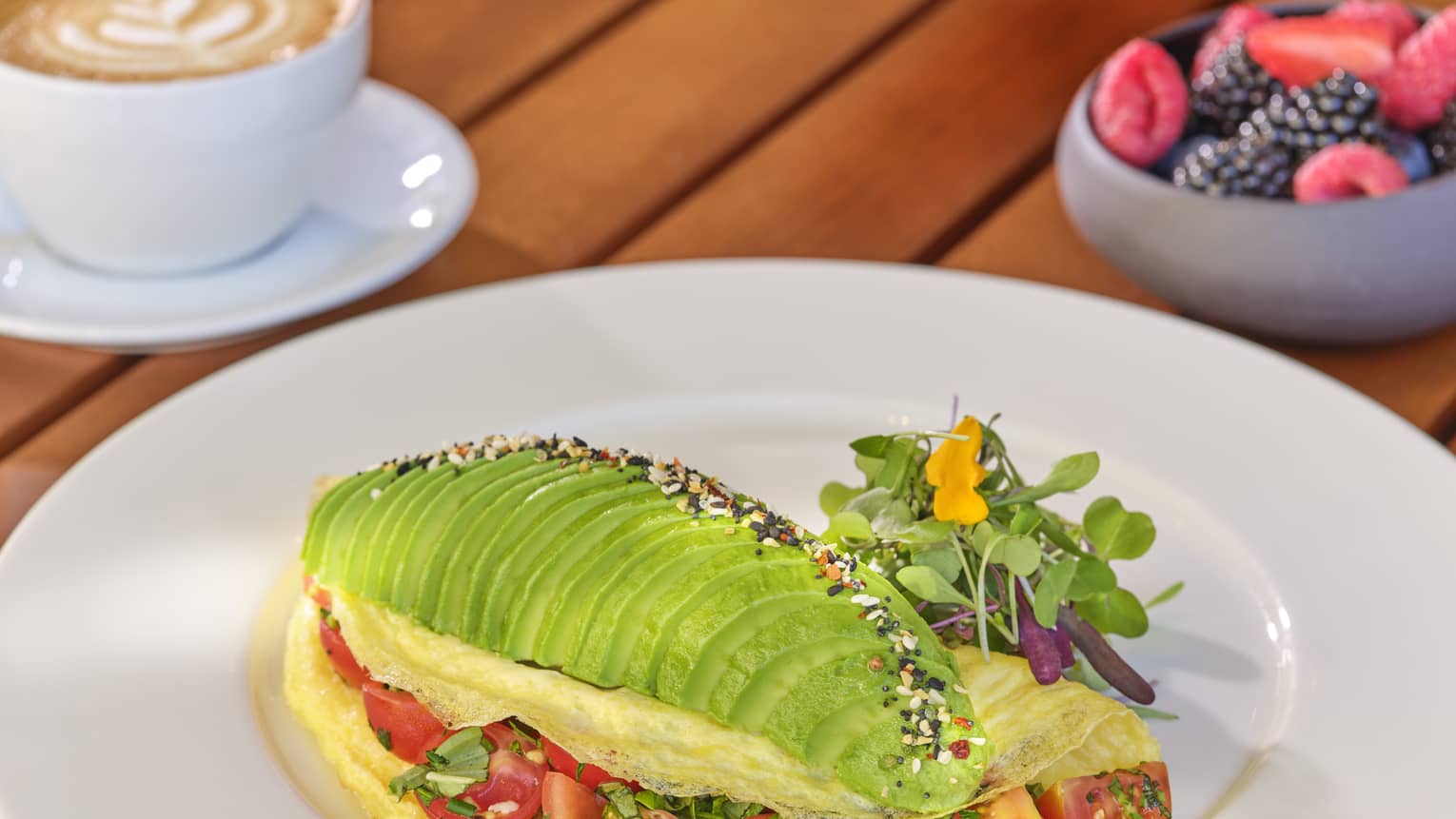 Omelette filled with cherry tomato halves, topped with row of sliced avocado and microgreen garnish on wood table beside latte and bowl of mixed berries