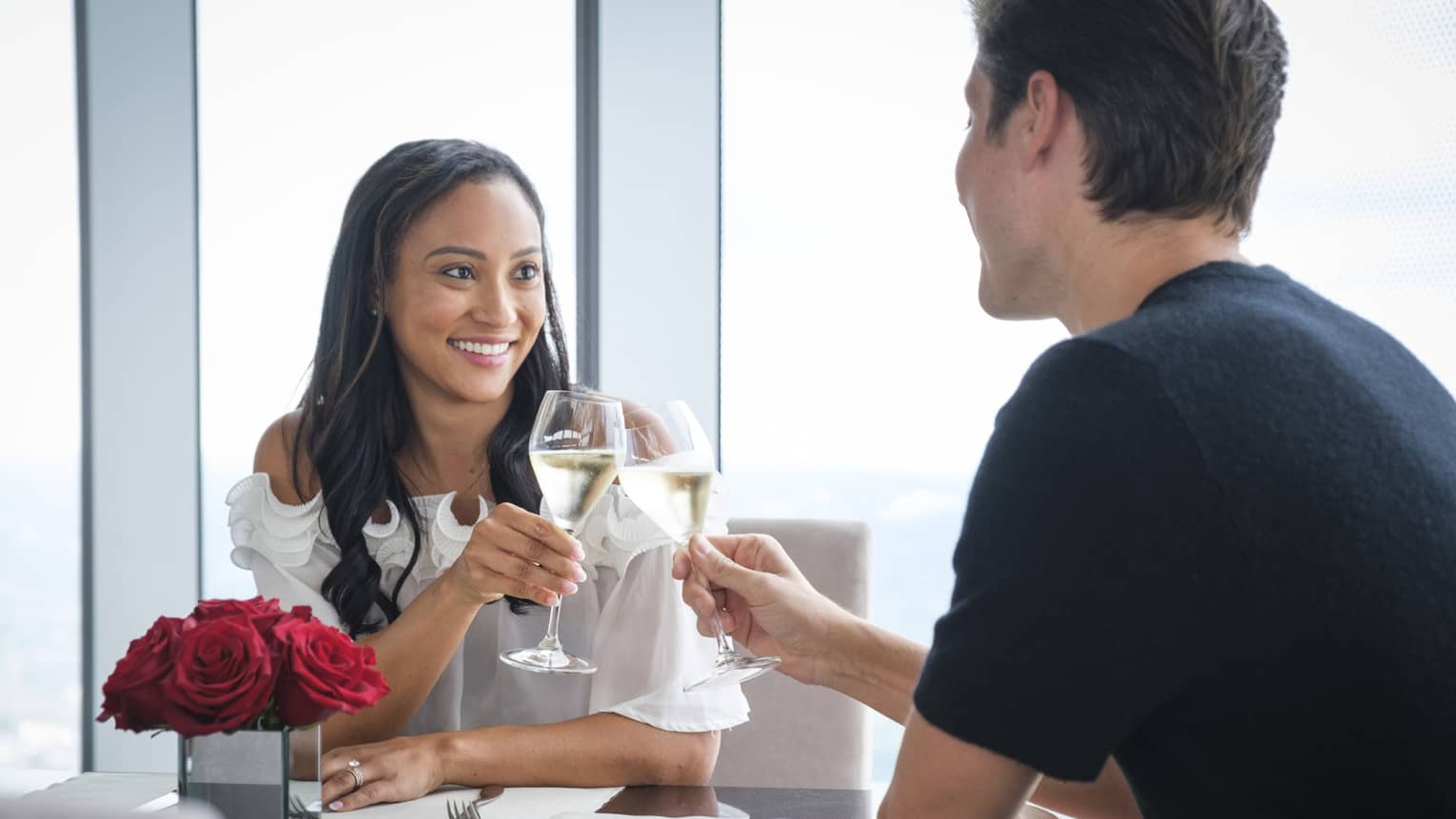 A man and woman celebrating with wine and red roses on a table near a large window.